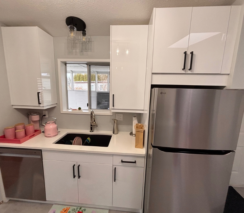 Bright kitchen renovated with white cabinets, a black sink and faucet by a window, pink countertop items, and a stainless refrigerator in the foreground with a ceiling light above.